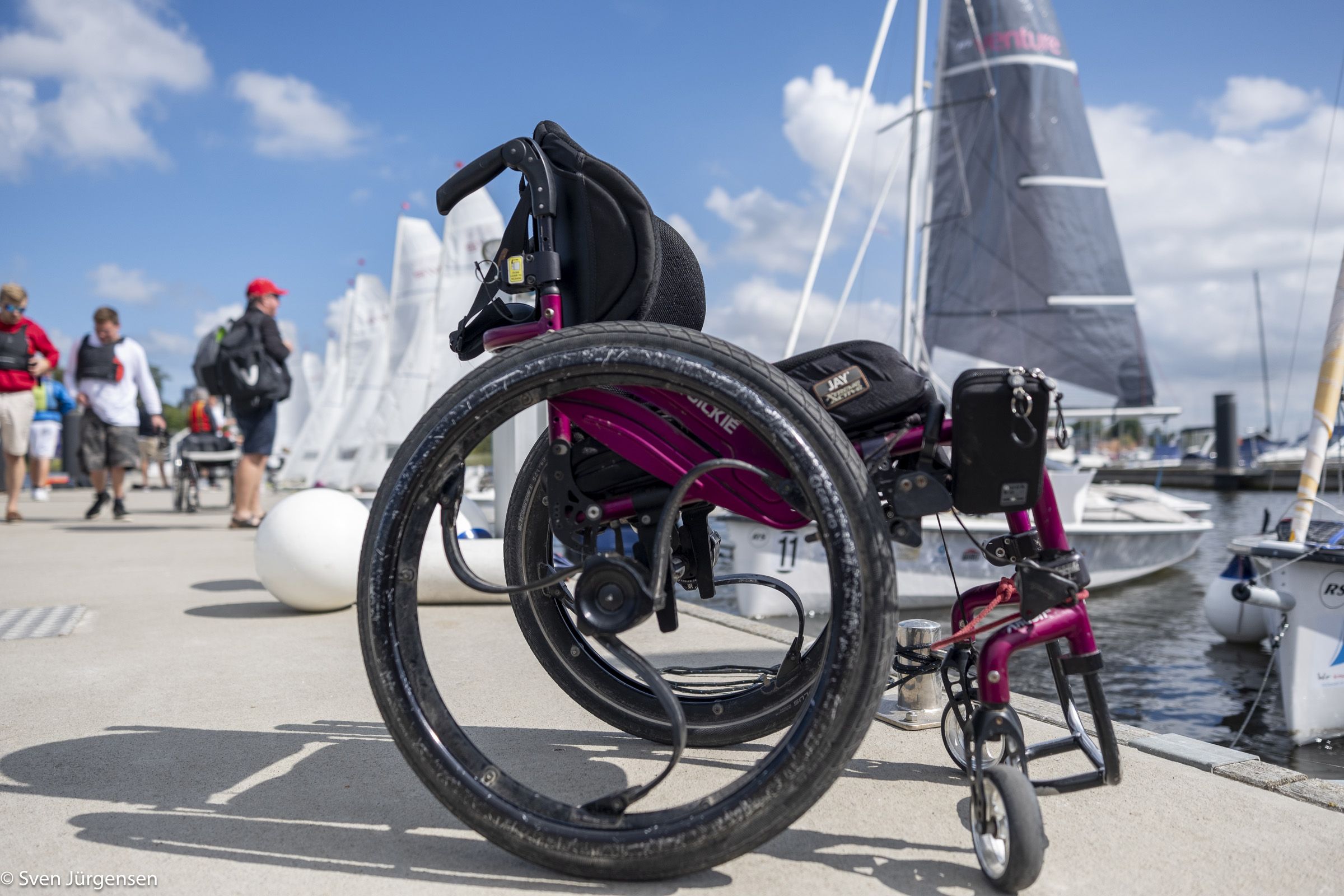 Rollstuhl im Vordergrund auf einem betonsteg mit einem Segelboot im Hintergrund