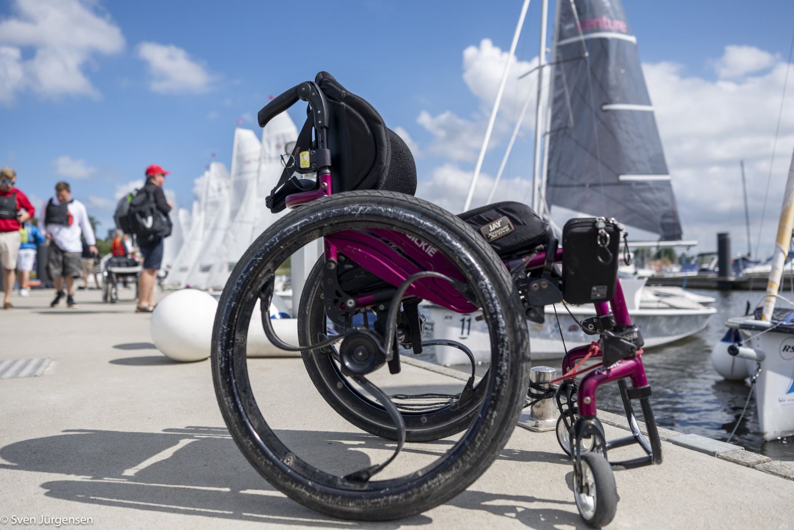 Rollstuhl im Vordergrund auf einem betonsteg mit Segelbooten im Hintergrund