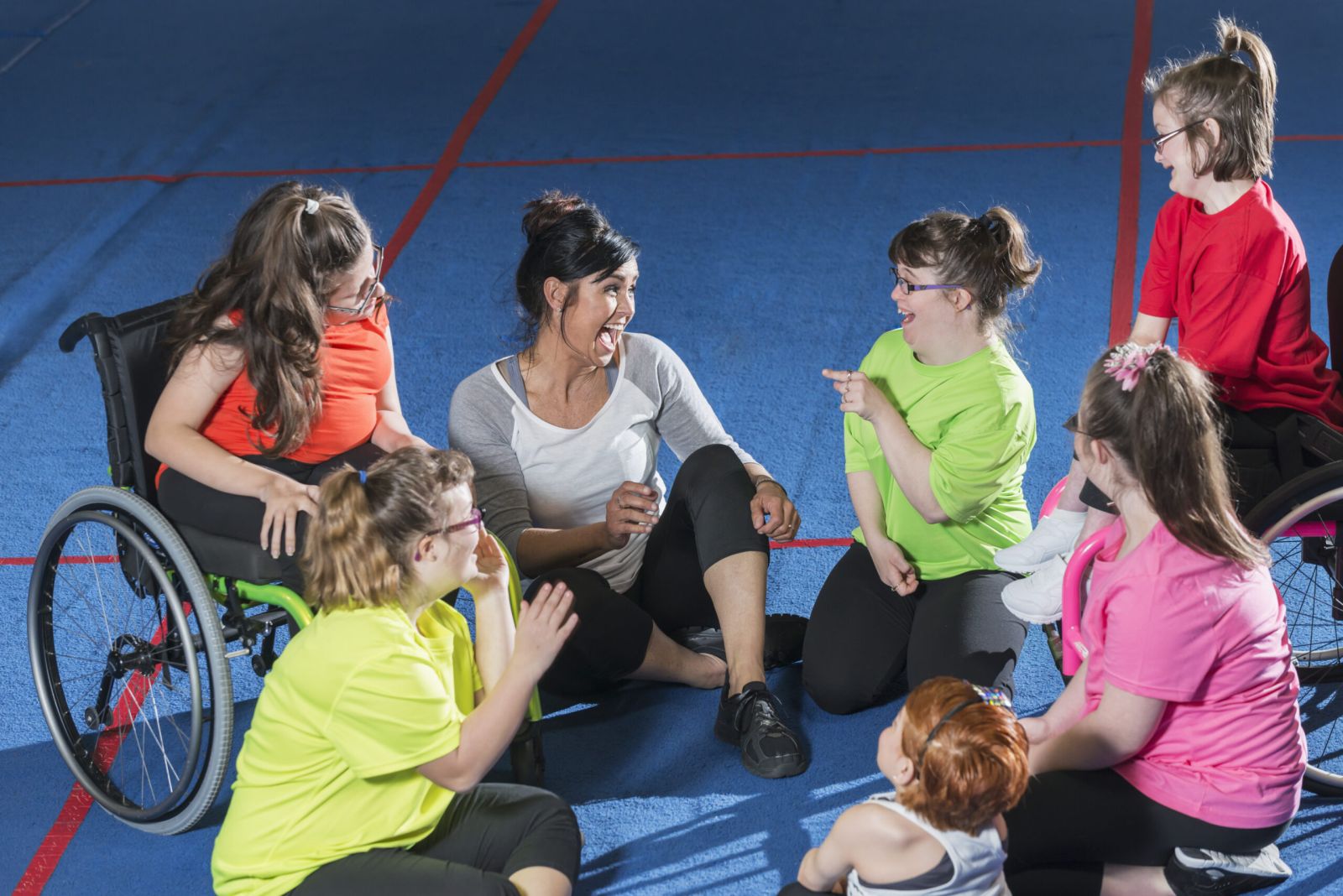 Ein Ausbilder sitzt mit einer Gruppe von Jugendlichen und jungen Frauen mit besonderen Bedürfnissen im Kreis auf dem Boden einer Sporthalle und spricht mit ihnen. Die Mädchen haben verschiedene geistige und körperliche Behinderungen, einschließlich Down-Syndrom und Morquio-Syndrom. Zwei der Jugendlichen sitzen im Rollstuhl.